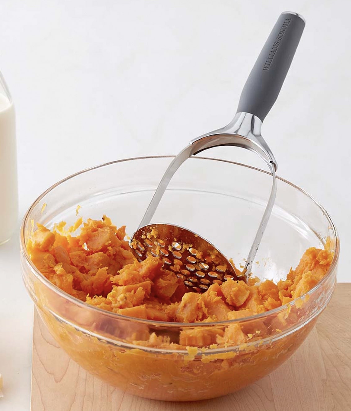 A metal potato masher is resting in a glass bowl filled with instant pot mashed potatoes made from sweet potatoes. A bottle of milk is visible in the background on the left.